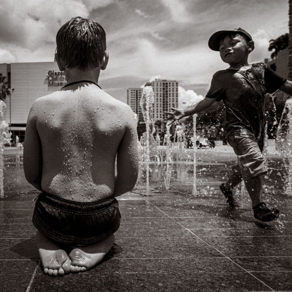 monochromatic child sitting on sidewalk as another kid splashes him with water