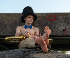 Barefoot shirtless boy wearing a top hat holding a saxophone.