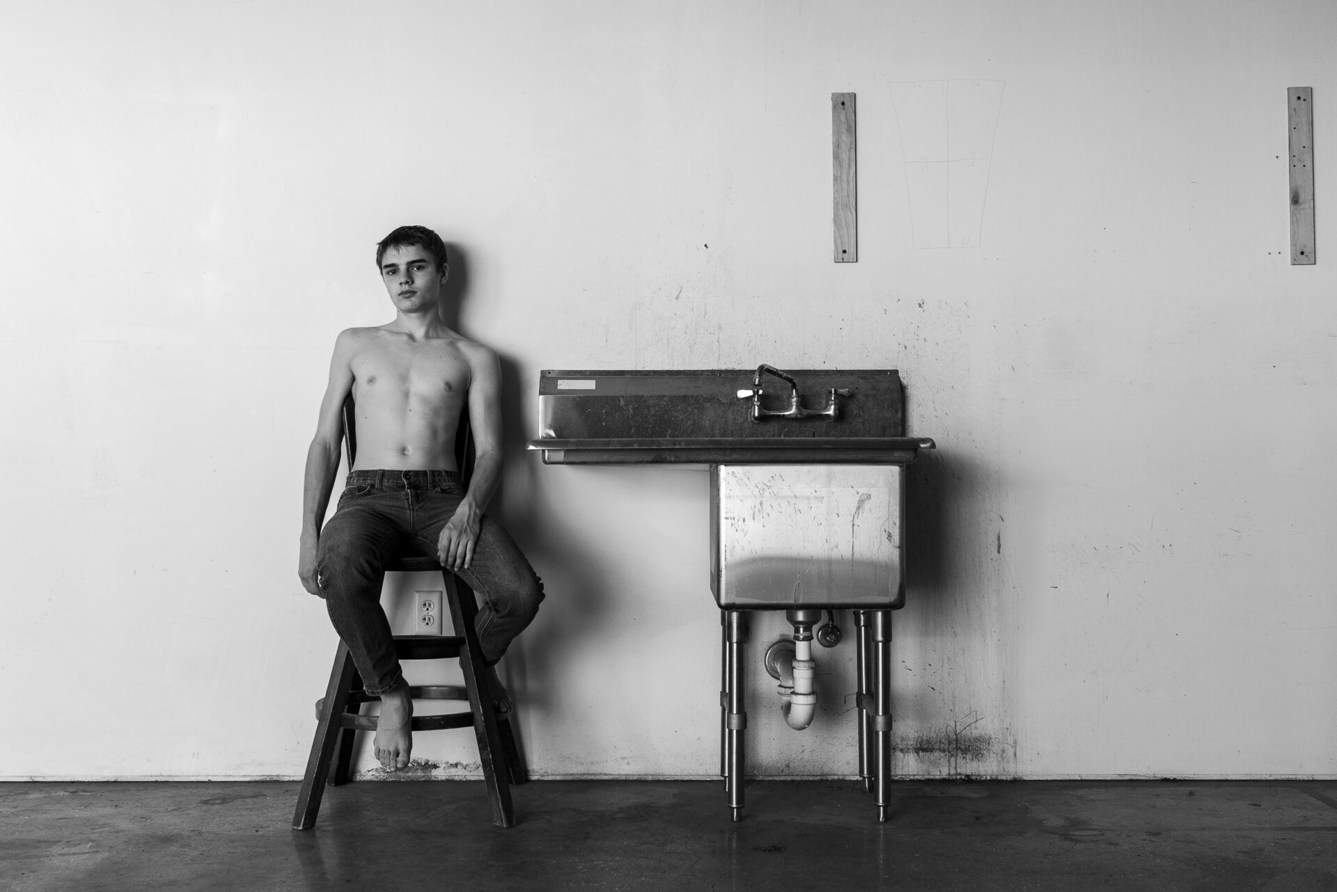 A shirtless boy in a black and white photo relaxes carelessly next to a stainless steel sink.