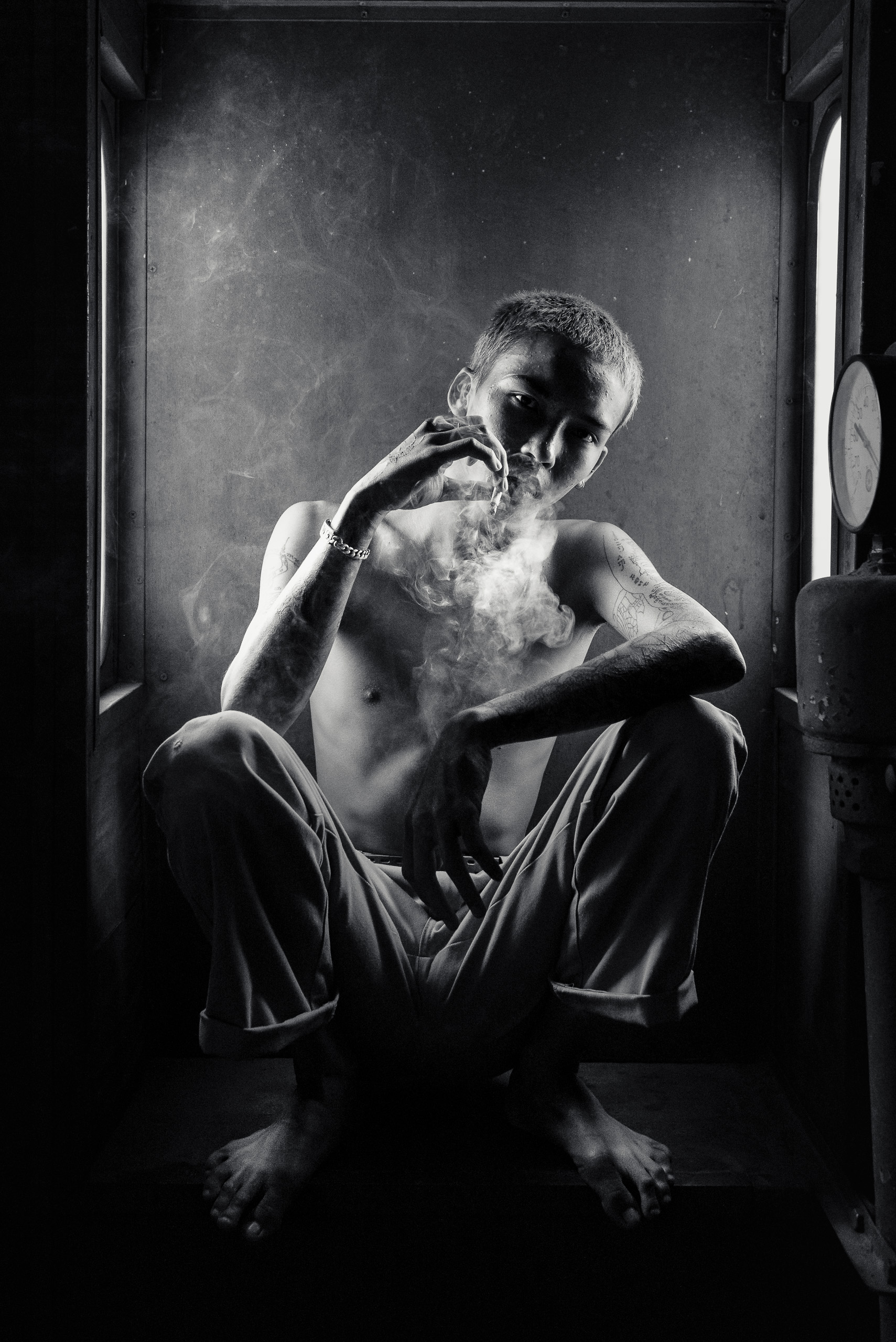 Fine art black and white portrait of a shirtless 17-year-old tattooed Thai boy sitting barefoot and smoking inside an abandoned army train in Bangkok, Thailand.
