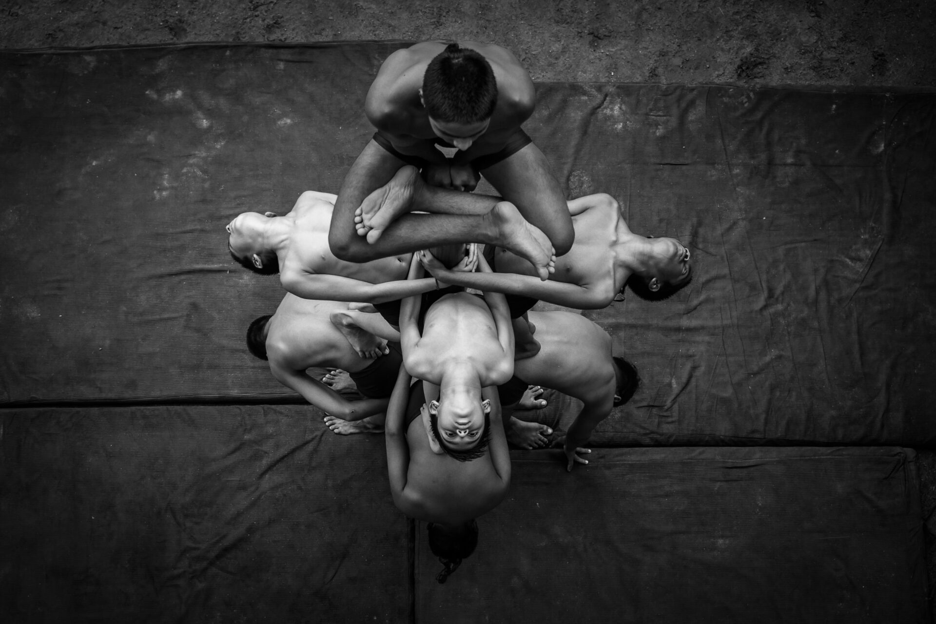 A high-angle black and white fine art photograph showing a group of young boys interlocked in a complex, symmetrical Mallakhamb human pyramid formation on training mats in Ahmedabad, India.