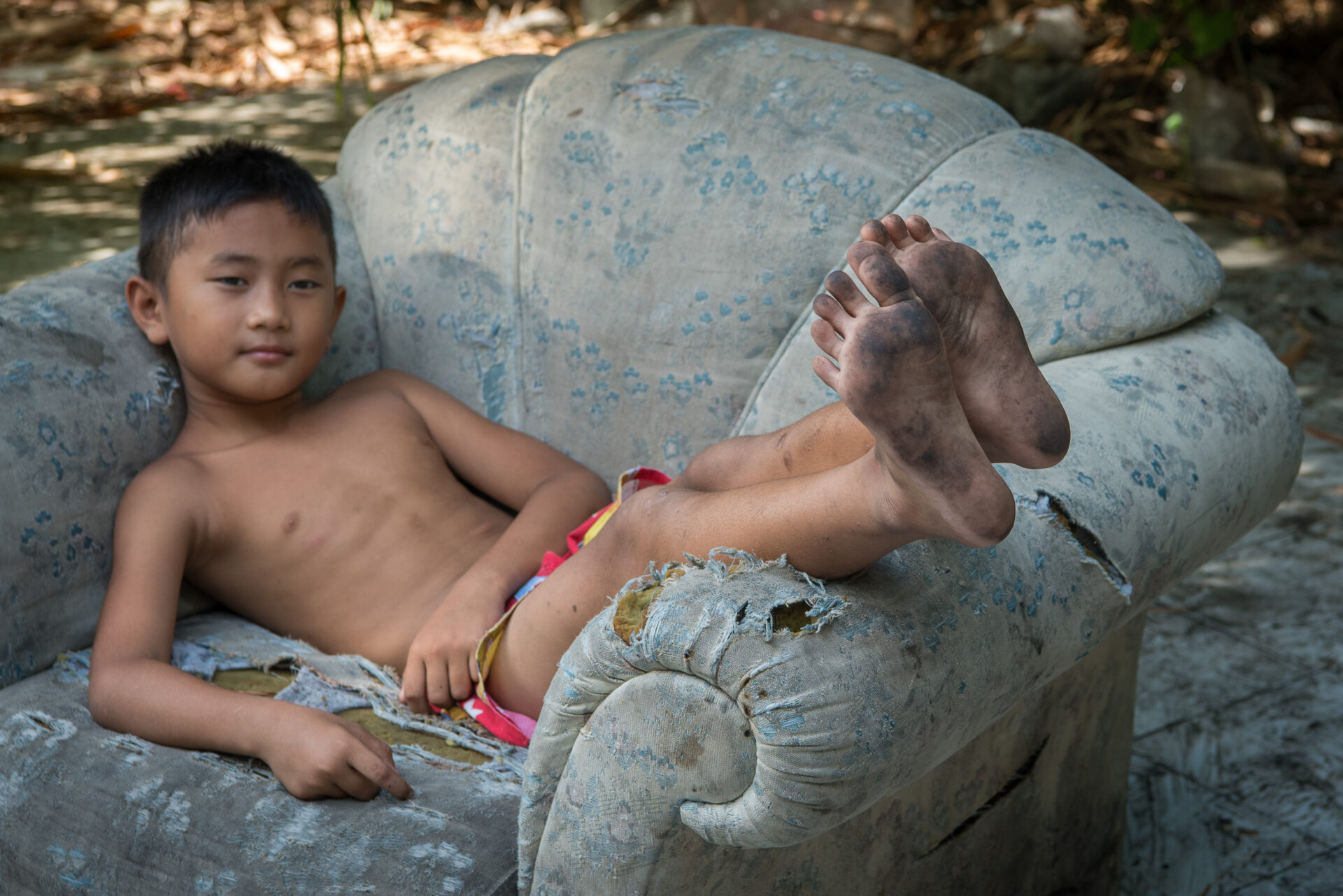 Young boy sitting on a worn armchair with dirty bare feet.