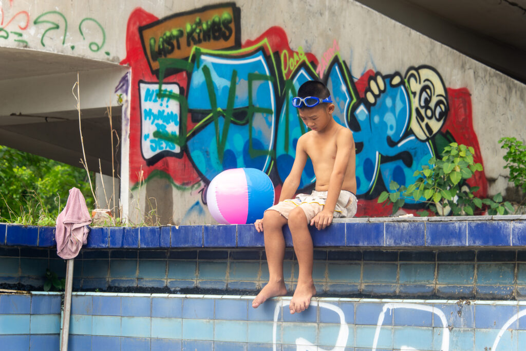 A young boy in swim goggles sitting thoughtfully on a concrete ledge next to vibrant graffiti and a beach ball at the Ghost Tower.