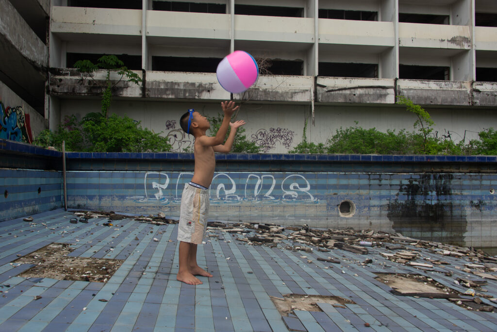 Wide shot of a boy playing with a pink balloon inside an abandoned rooftop pool structure at the Ghost Tower in Chiang Mai.