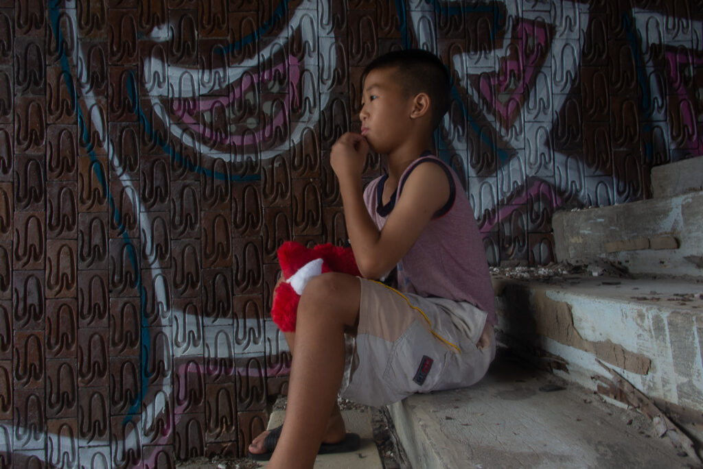 Profile portrait of a young boy sitting cross-legged on concrete steps in front of a dark wall with graphic red graffiti.